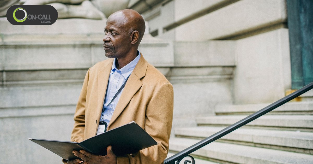 A man in a tan overcoat holds an open black folder on the stone steps of a building with a metal handrail