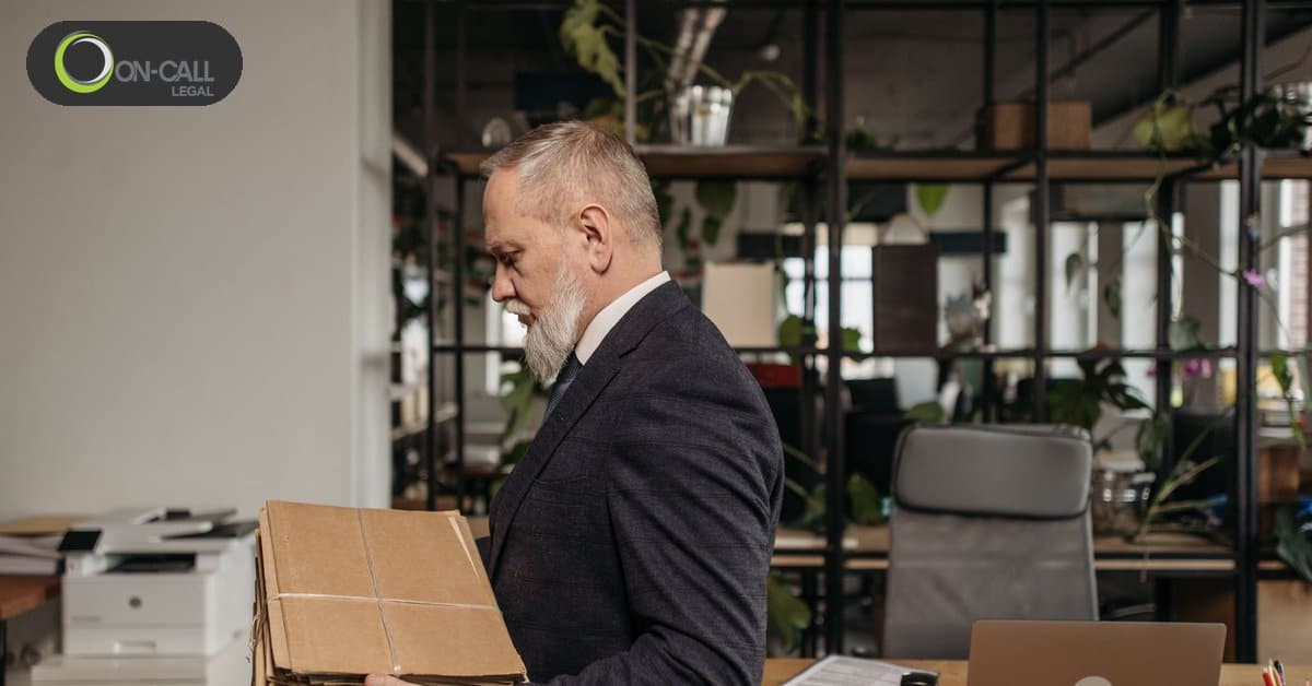 A gray-haired man in a dark suit holds a flat cardboard package next to a printer in an open office