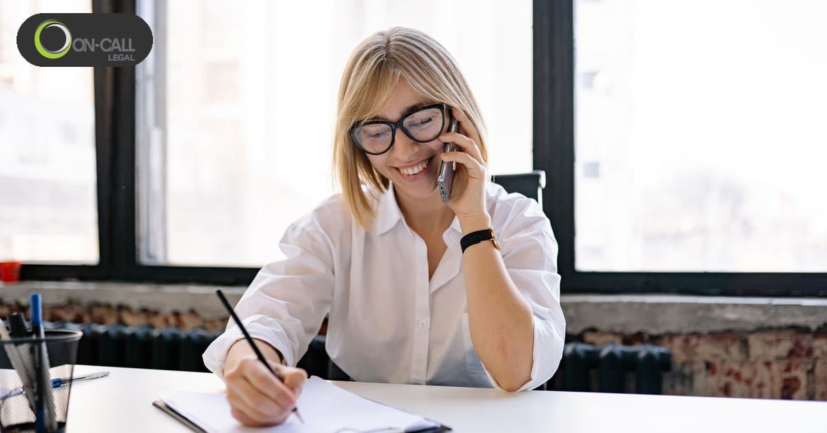 A smiling woman in glasses talks on the phone and writes notes on a clipboard at a white desk