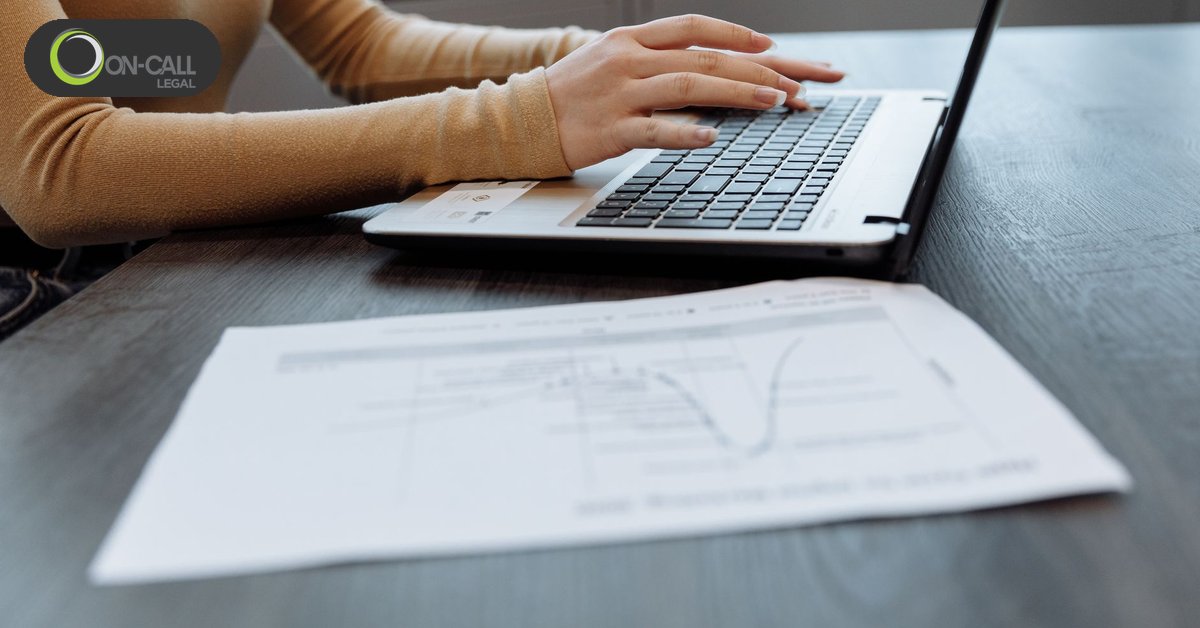 A woman types on a laptop at a dark table with a printed page lying in front of the keyboard