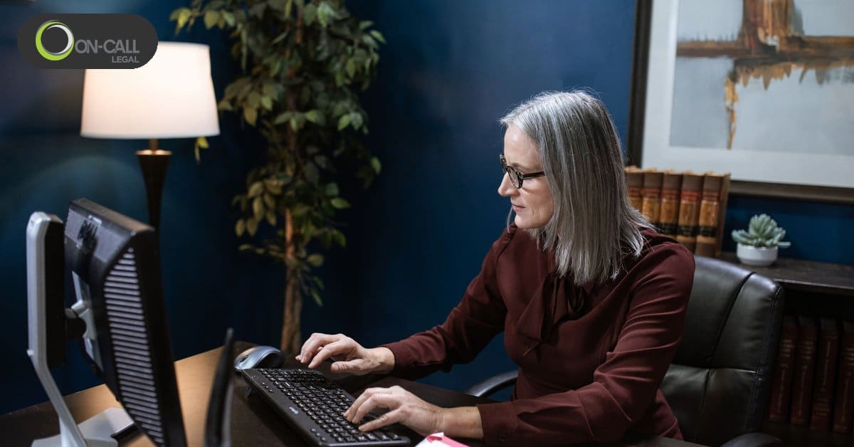A woman with gray hair and glasses types at a desktop computer in an office with a dark blue wall