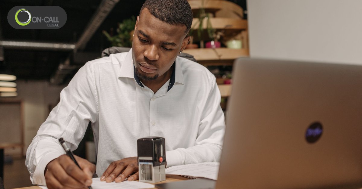 A man in a white shirt writes on a document at a desk next to a self-inking stamp and a laptop