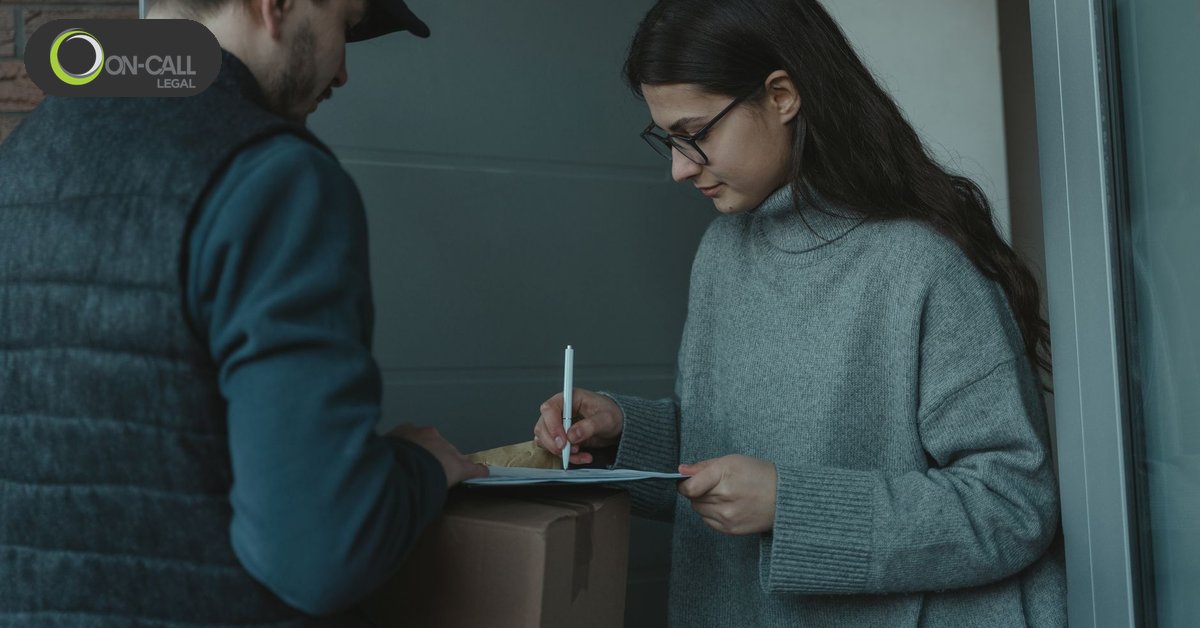 A woman in a grey sweater signs a slip at her door while a man in a cap holds a parcel