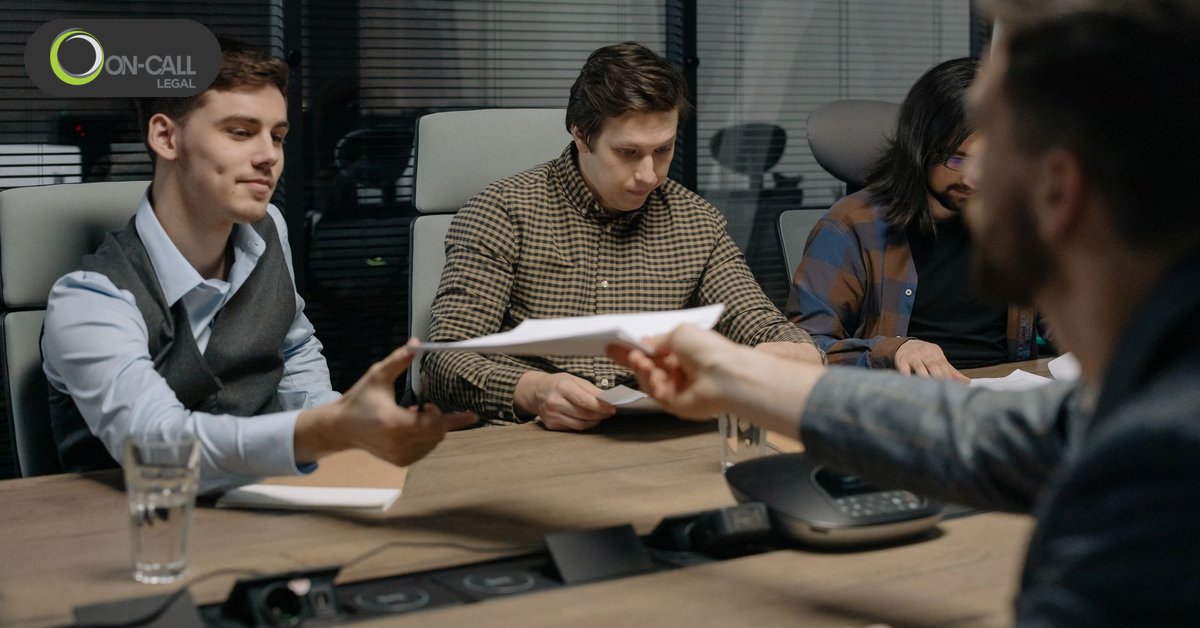 A man hands a set of papers across a conference table to a seated man in a vest during a meeting