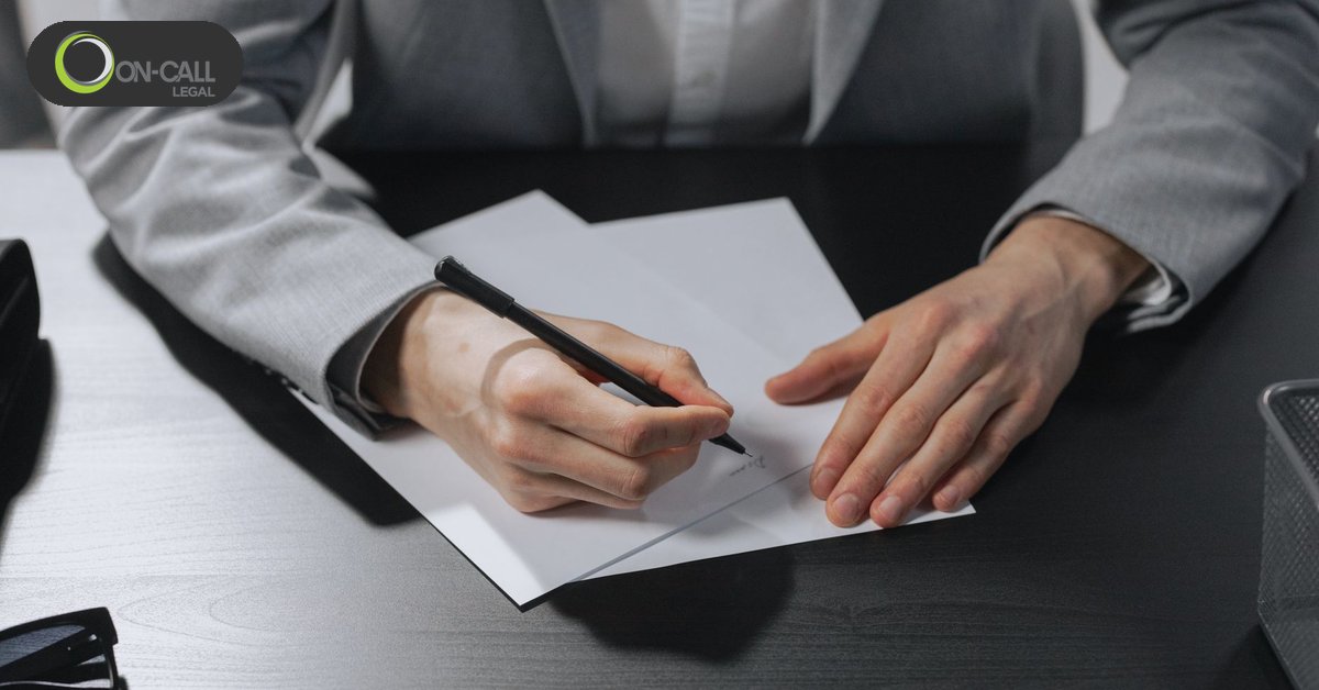 A person in a grey blazer signs a sheet of paper at a dark desk, the other hand holding it flat