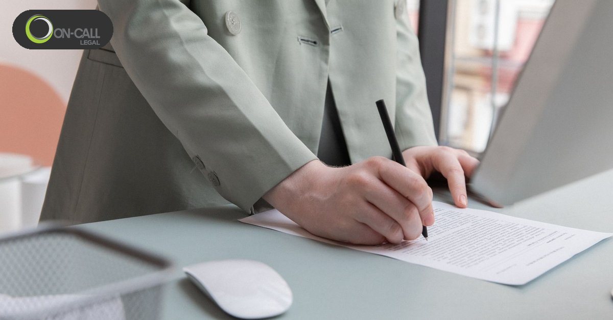 A person in a sage green blazer signs a one-page document at an office desk