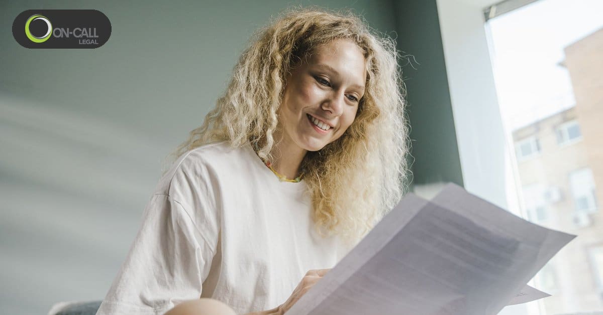 A smiling woman with curly blonde hair reads a printed letter while sitting by a window at home