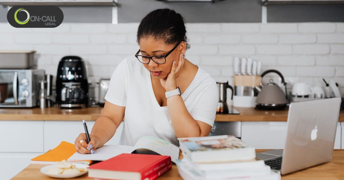 A woman in glasses takes notes at a kitchen table next to a laptop and a stack of books