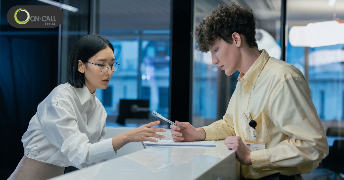 A receptionist in a white shirt talks with a visitor across an office reception counter with papers on it